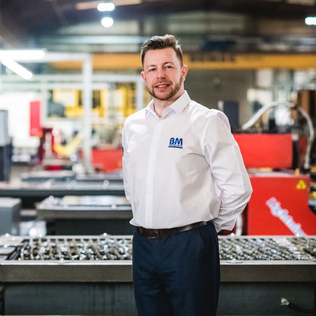 A BM Catalysts employee is stood in the factory next to heavy machinery. He is wearing a BM branded shirt and is smiling.