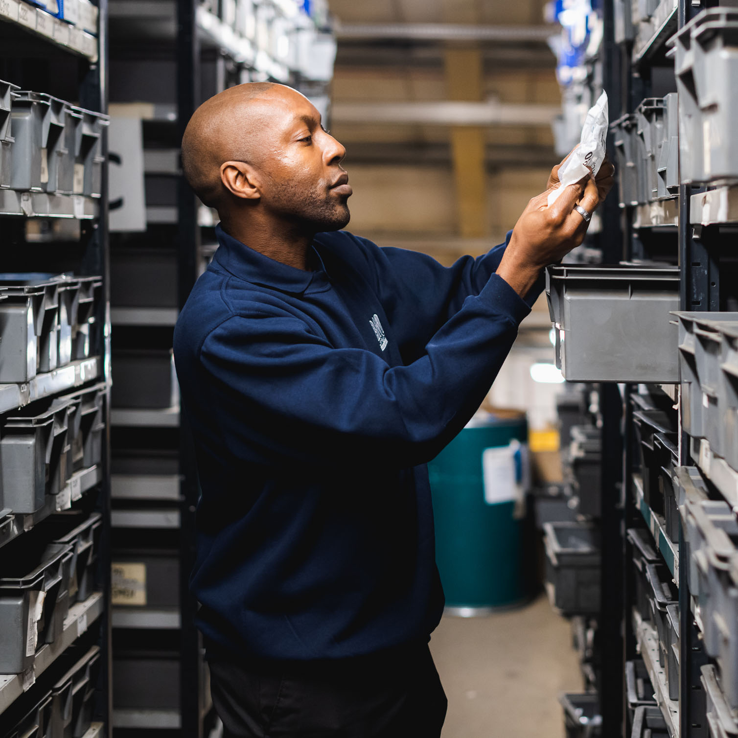 A man in a warehouse is looking at a packaged product for a vehicle part which he pulled out from a storage contain draw. 